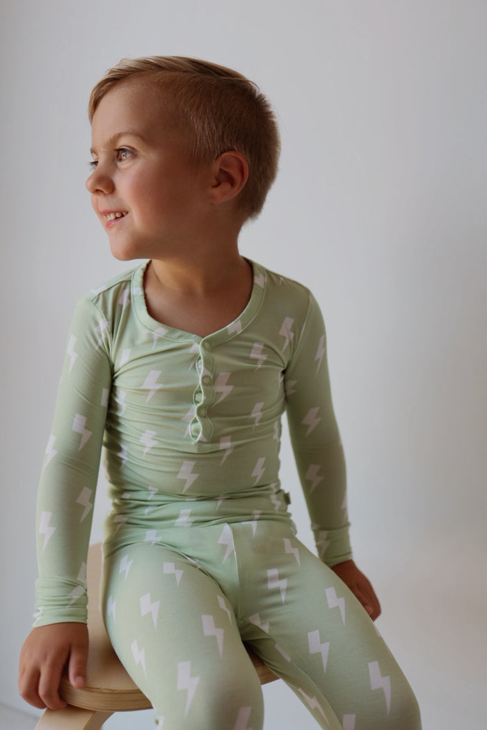 A young child with short light brown hair, smiling and sitting on a wooden stool, wears forever french baby’s Bamboo Two Piece Pajamas | Flash—light green pajamas with white lightning bolts—against a plain background.
