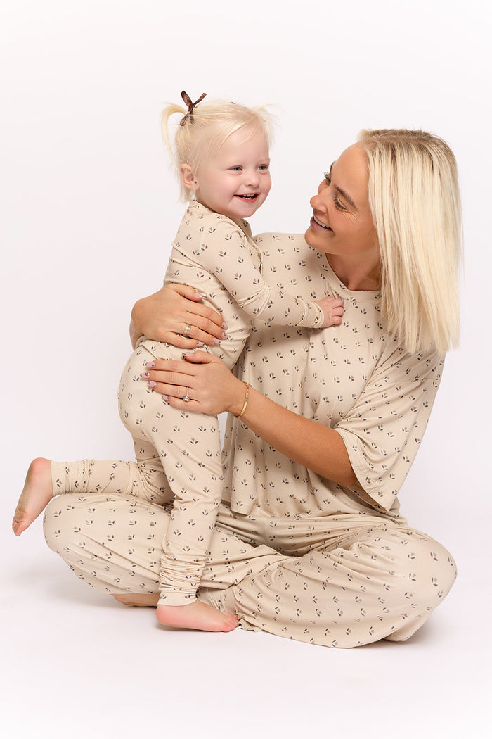 A smiling woman and happy toddler sit on the floor in matching forever french baby Bamboo Two Piece Pajamas | Flora—beige bamboo sleepwear with delicate patterns, ideal for hypoallergenic kids. The background is plain white.