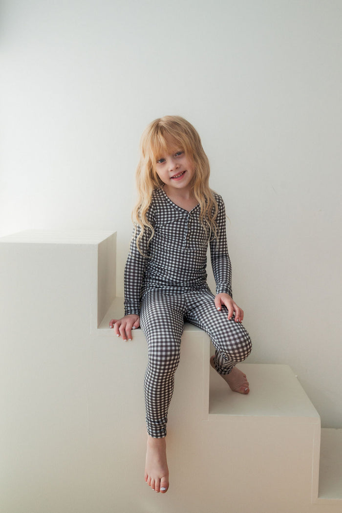A young child with long blonde hair, barefoot and smiling at the camera, wears forever french baby’s Bamboo Two Piece Pajamas in Green Gingham while sitting on white block steps against a plain white background.