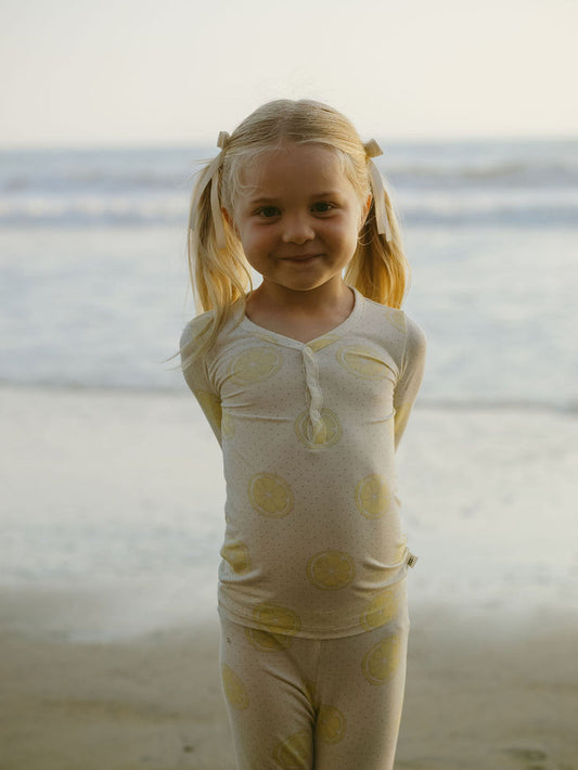 A young girl with blonde pigtails stands on a beach wearing forever french baby’s Bamboo Two Piece Pajamas in Lemon Drop, featuring yellow citrus prints. She smiles with her hands behind her back as the softly blurred ocean and sky create a dreamy backdrop.