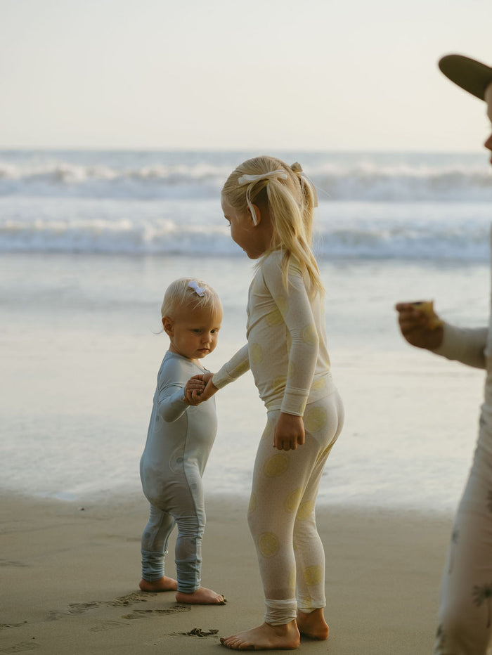 Two young children wearing forever french baby Bamboo Two Piece Pajamas in Lemon Drop stand barefoot by the water, holding hands as the older gently guides the younger. Waves and soft sunlight create a peaceful, dreamy beach scene.