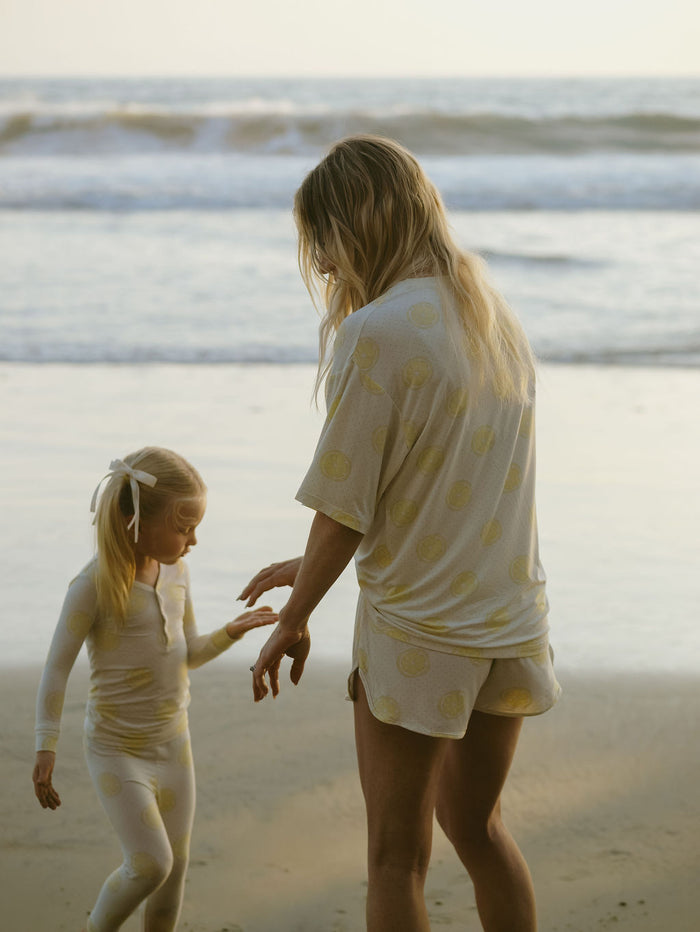 An adult and a young girl wear matching Lemon Drop bamboo pajamas by forever french baby on a sandy beach by the ocean. The hypoallergenic kids' Bamboo Two Piece Pajamas provide comfort and style for little ones.