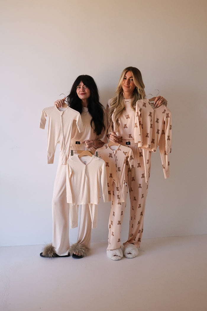 Two women in matching cream-colored Bamboo Zip Pajamas by forever french baby hold up children’s pajama sets—one solid, one with a small teddy bear print. Both wear slippers and stand against a plain background.