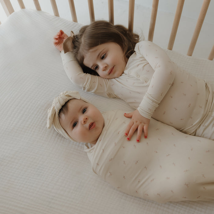 Two children rest in a crib: an older child in pajamas gently touches a baby wrapped in the forever french baby Bamboo Swaddle | Lucy with a matching headband, both lying on a soft, hypo-allergenic blanket.