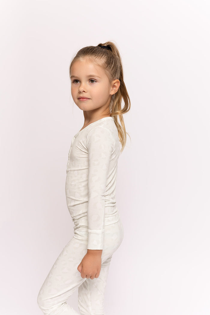 A young girl with long, light brown hair in a ponytail stands sideways, wearing the forever french baby Bamboo Two Piece Pajamas | Meow. The light-colored, breathable set features a long-sleeve top and pants for all-day comfort against a white background.