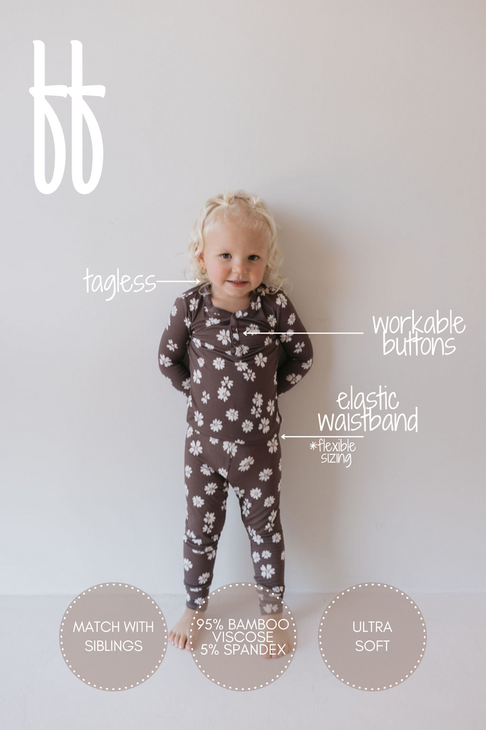 A toddler with blond hair stands against a light background wearing the "Midnight Meadow" two-piece pajamas from forever french baby, featuring a brown onesie embellished with white flower patterns. These hypoallergenic pajamas boast workable buttons, a tagless design, and an elastic waistband, crafted from 95% breathable bamboo clothing and 5% spandex.