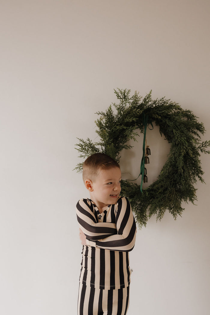 A young boy in Minty Cherries Bamboo Two Piece Pajamas | Midnight Stripe stands smiling with arms crossed before a plain wall, a green holiday wreath hanging behind him.