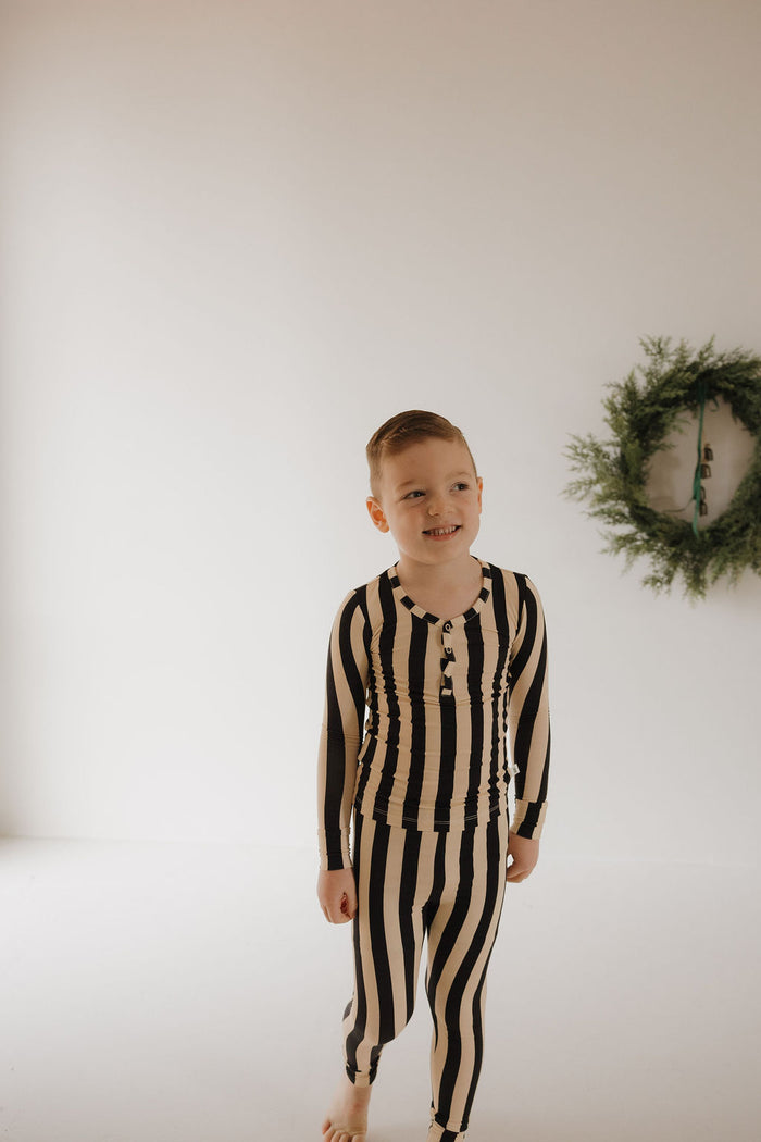 A young boy smiles barefoot in a white room, looking left in Minty Cherries' Bamboo Two Piece Pajamas | Midnight Stripe. A green wreath on the wall adds a festive touch to his soft, breathable sleepwear.