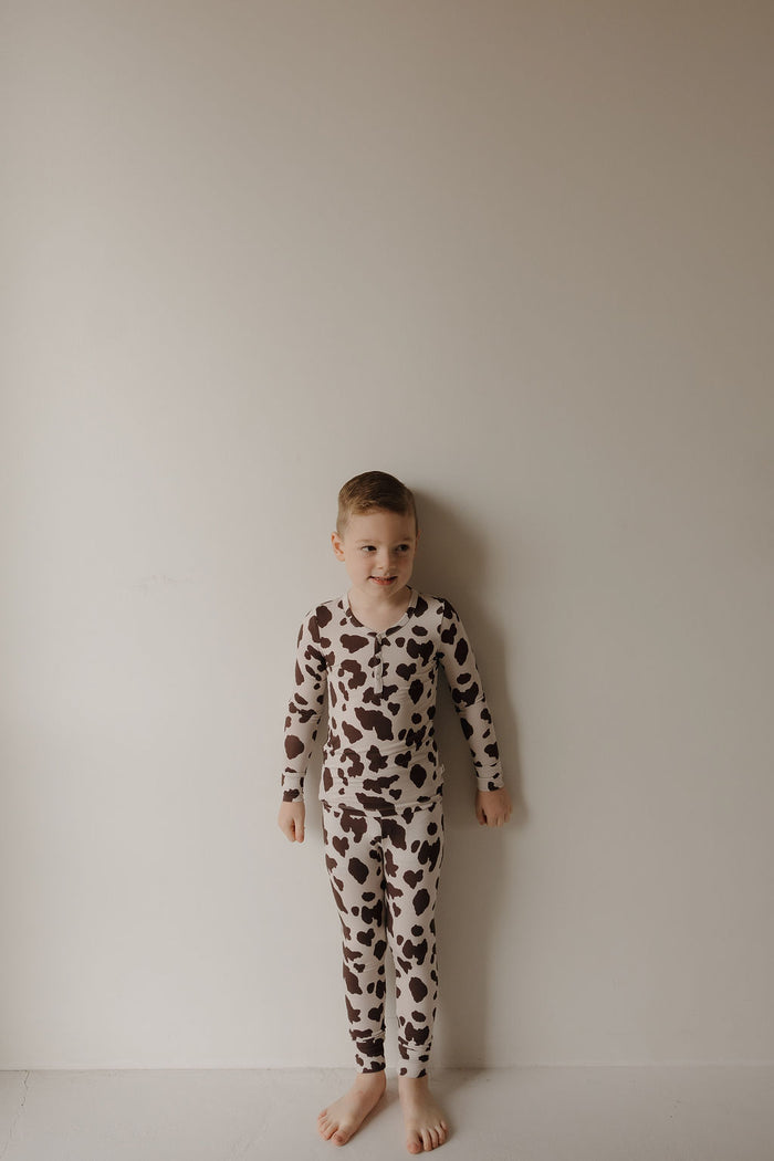 A young boy stands barefoot against a light wall, wearing forever french baby’s Bamboo Two Piece Pajamas | Moo!—long-sleeved with a brown and white cow print—showing off the soft, breathable comfort of bamboo kids sleepwear.