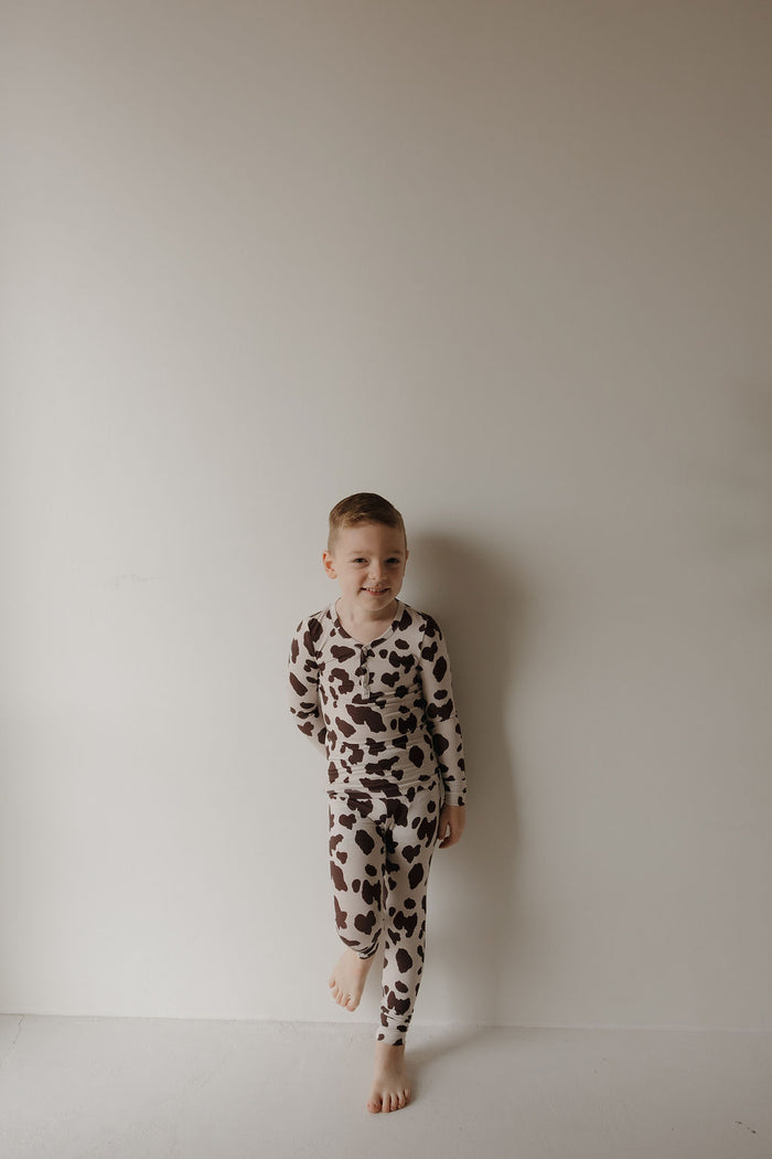 A young boy stands barefoot against a light wall, smiling in matching brown and white Bamboo Two Piece Pajamas | Moo! by forever french baby. His hypo-allergenic kids bamboo sleepwear is comfy, stylish, and perfect for bedtime. One knee is slightly bent.