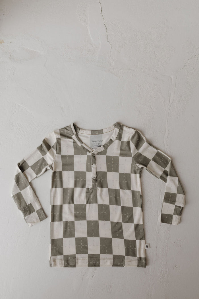 The Bamboo Two Piece Pajamas in Oat & Sage by forever french baby features a buttoned neckline, offering breathable comfort and style. The long-sleeved, checkered top with green and white squares is laid flat on a textured white surface.