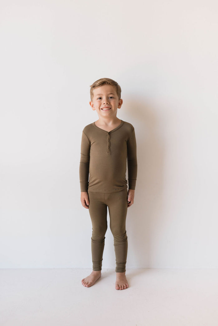 A young boy stands barefoot against a plain white background, smiling and wearing matching olive green long-sleeve top and pants from Forever French Baby's Bamboo Two Piece Pajamas. His hypo-allergenic sleepwear is made from bamboo fabric. His hands are relaxed at his sides, and he has short, light-colored hair.