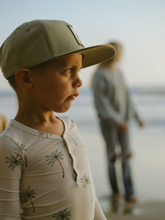 A young boy in forever french baby's Bamboo Two Piece Pajamas | Palm Tree stands on the beach, wearing a cap and looking to the side. His breathable sleepwear keeps him cool, while another person is visible near the shoreline.
