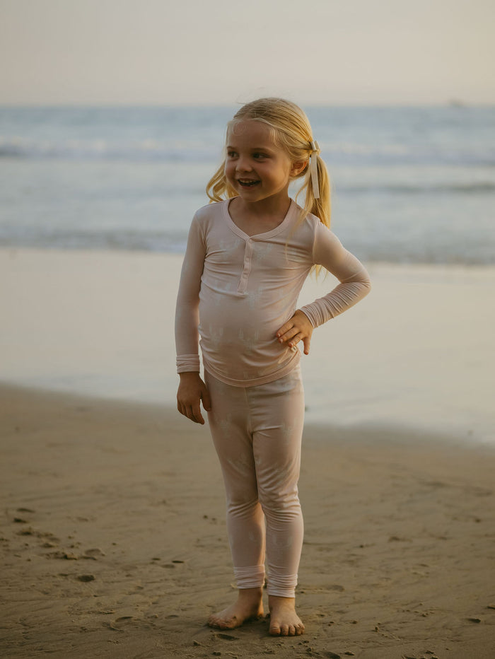 A smiling blonde girl in pigtails poses barefoot on the beach, ocean behind her, wearing forever french baby Bamboo Two Piece Pajamas | Lobster—breathable, comfy children's sleepwear.