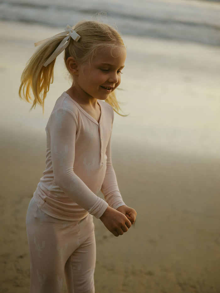 A young blonde girl in pigtails wears forever french baby's Bamboo Two Piece Pajamas | Lobster—light pink, hypoallergenic kids sleepwear with a subtle pattern—while smiling and playing on the beach at sunset.