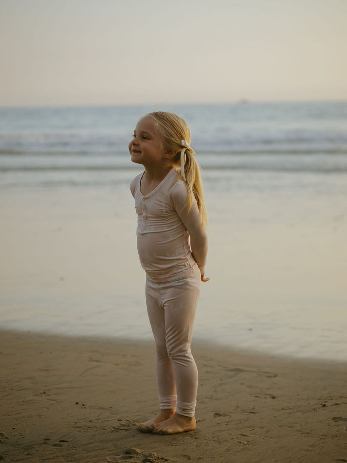 A young blonde girl stands barefoot on the beach, smiling with her hands behind her back. She wears forever french baby’s Bamboo Two Piece Pajamas in Lobster as the calm sea and sky form a peaceful backdrop.