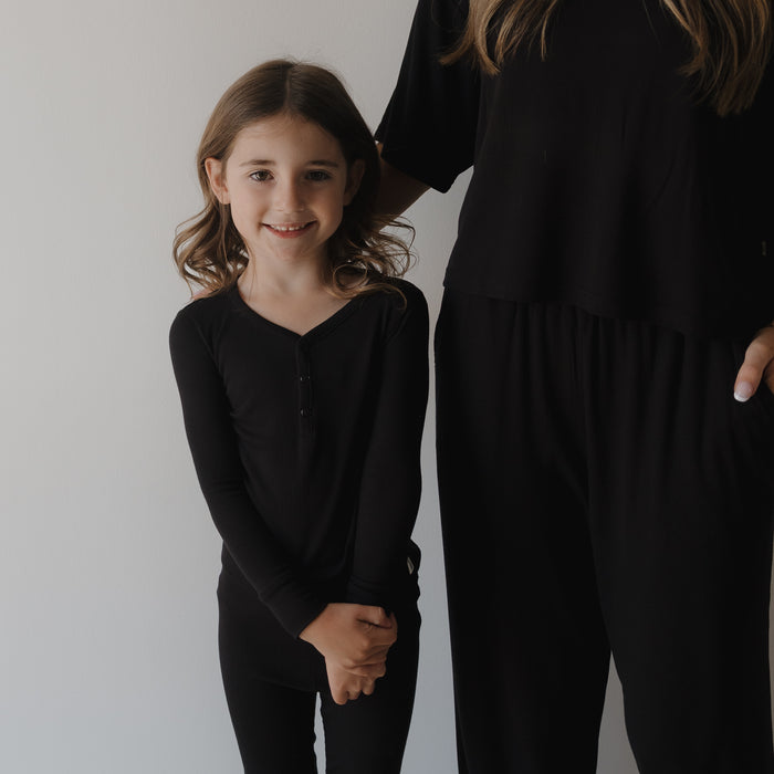 A young girl with wavy hair, smiling in forever french baby Bamboo Two Piece Pajamas | Ribbed Black, stands next to a partially visible adult in loose black clothing against a plain light background.
