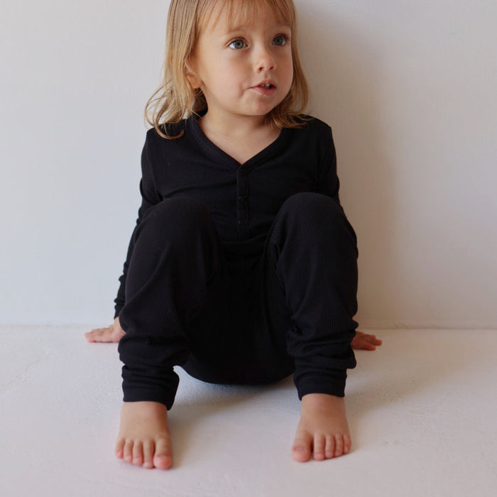 A young child with shoulder-length blonde hair, dressed in Forever French Baby's Bamboo Two-Piece Pajamas in Black Ribbed, is sitting against a white wall. The child's legs are bent, feet flat on the ground, and hands resting between their legs. The child gazes into the distance with a thoughtful expression.