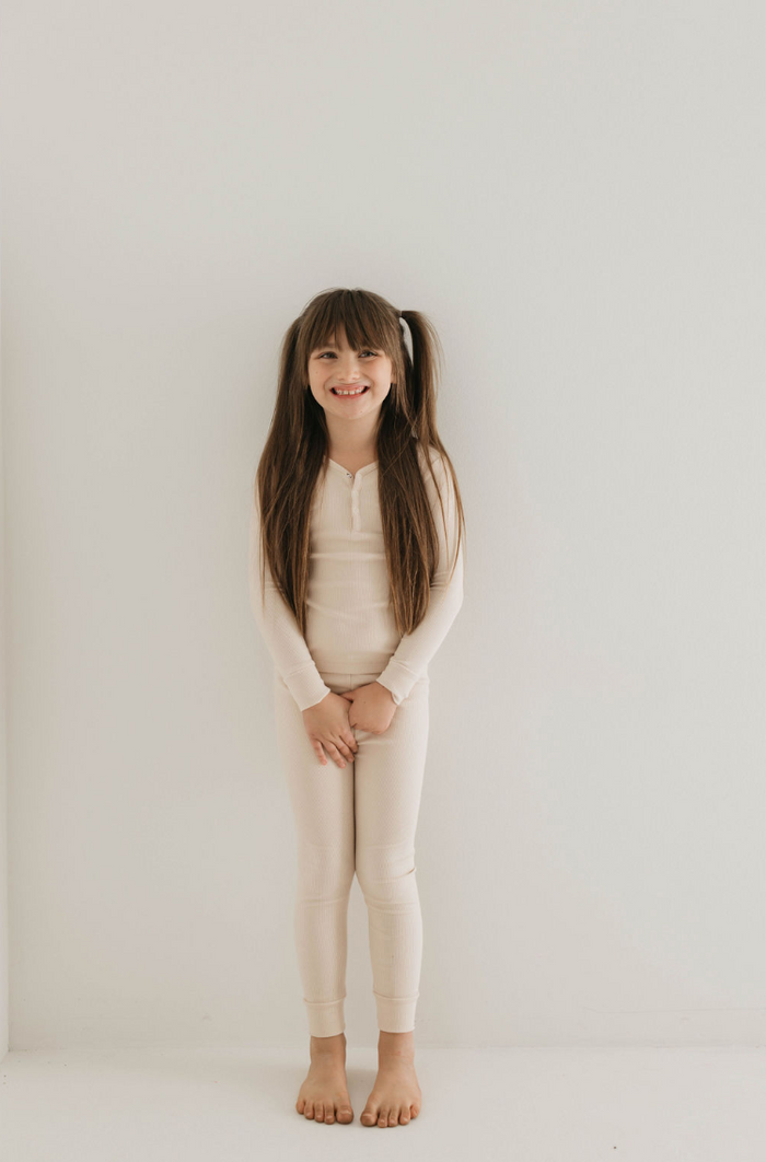 A young girl with long brown hair in pigtails stands smiling against a plain white background. She is wearing the "Two Piece Bamboo Pajamas | Ribbed Cream" by forever french baby, made from sustainable bamboo fabric and featuring matching leggings. Her ensemble looks as cozy as it is eco-friendly. With hands loosely clasped in front of her, she’s barefoot and adorable.