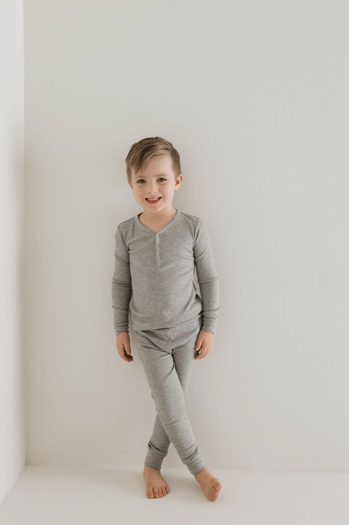 A young boy stands barefoot against a white wall, wearing Bamboo Two Piece Pajamas in Ribbed Grey by forever french baby. He smiles at the camera, with his right hand resting by his side and his left hand slightly behind his back, showcasing the comfort and style of eco-friendly kids sleepwear.