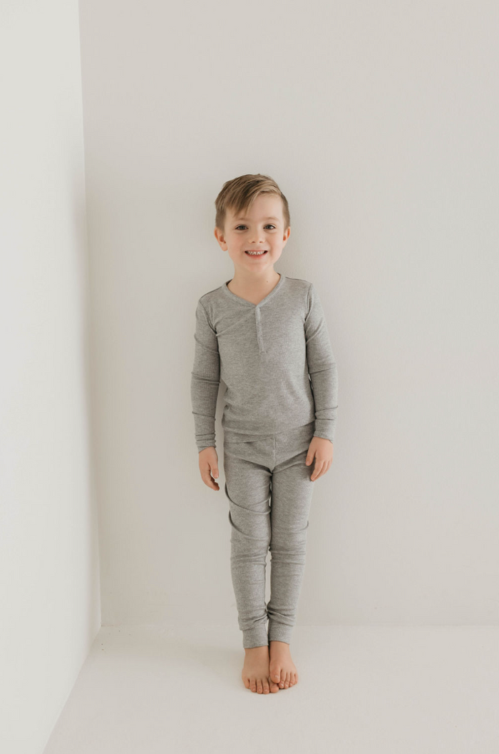 A young child stands barefoot against a white wall, smiling at the camera. Dressed in forever french baby's Bamboo Two Piece Pajamas in ribbed grey, the child radiates comfort and joy. This eco-friendly kids sleepwear enhances the minimalistic and bright atmosphere, emphasizing sustainability and style.