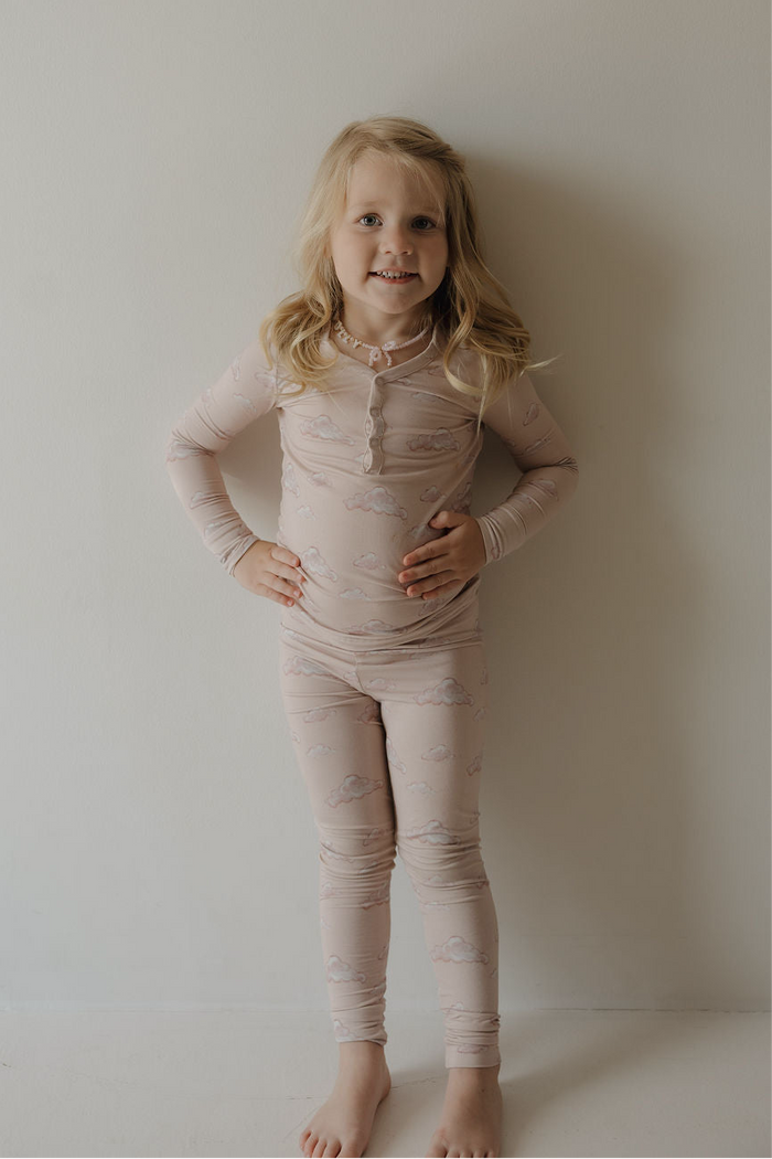 A young child with long blonde hair, barefoot against a light wall, smiles with hands on hips wearing Kendy 3’s Bamboo Two Piece Pajamas | Ruby—enjoying the breathable comfort of these matching pink cloud-patterned pajamas.