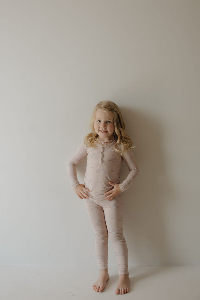 A young child with blonde hair stands barefoot against a light background, smiling in Kendy 3’s Bamboo Two Piece Pajamas | Ruby, featuring a subtle pattern and made from breathable bamboo fabric.
