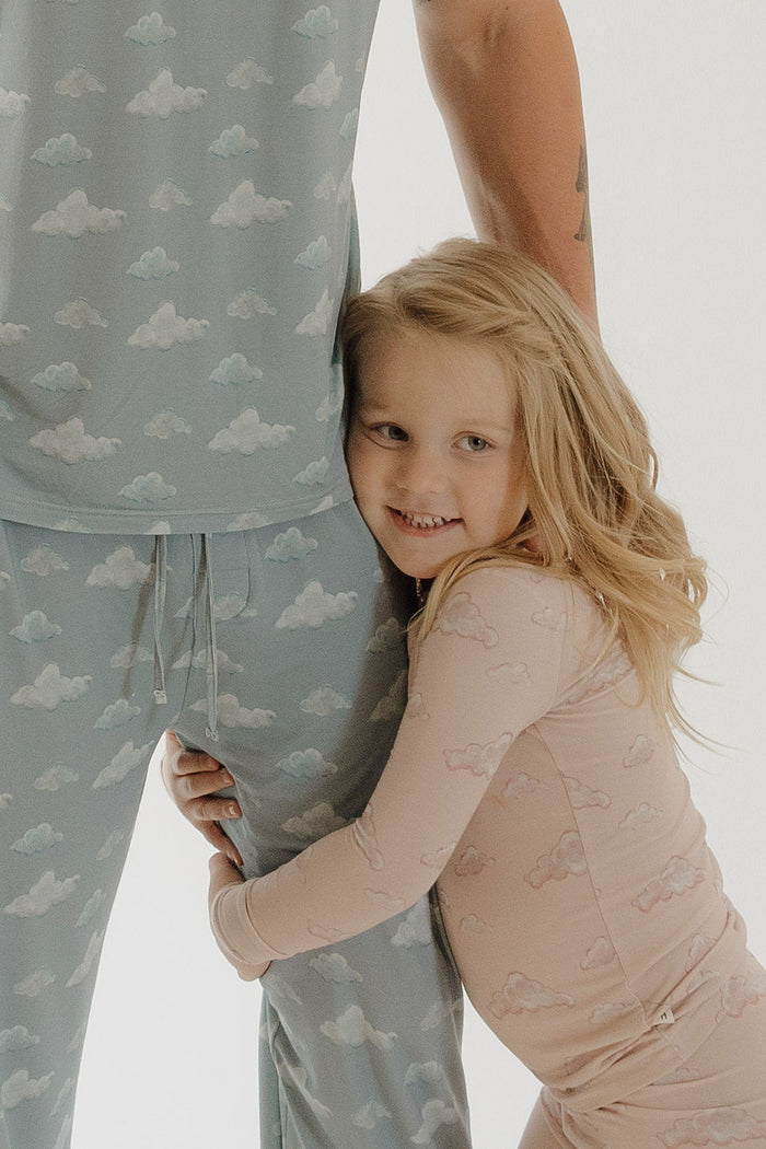 A smiling young girl wears Kendy 3’s Bamboo Two Piece Pajamas | Ruby in pink with a cloud pattern, hugging an adult’s leg in matching blue pajamas, both posed against a plain white background.