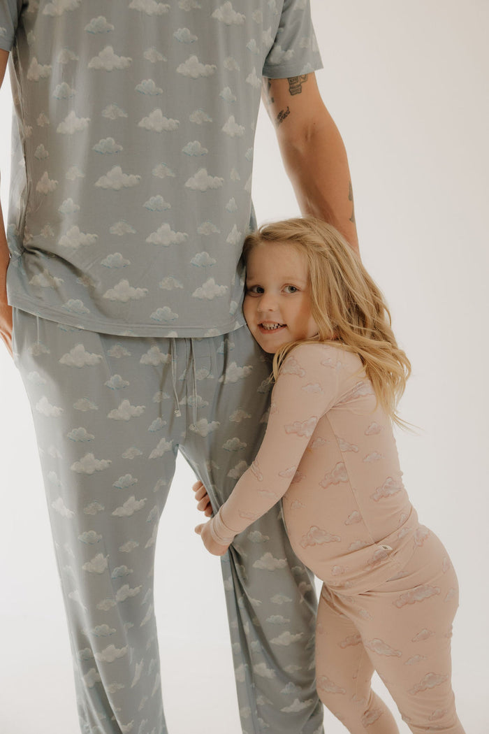 A young child wears Kendy 3's Bamboo Two Piece Pajamas | Ruby in pink cloud print, hugging the leg of an adult in matching blue sleepwear. The soft, breathable fabric stands out against a light, minimal background.