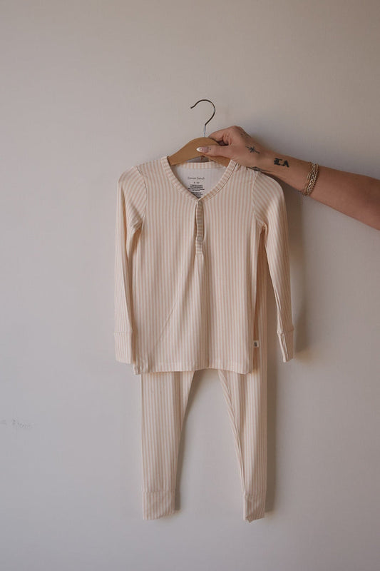 A hand displays forever french baby’s Bamboo Two Piece Pajamas in Sadie Stripe—light cream and beige striped, soft, and hypo-allergenic sleepwear for kids—against a plain wall on a wooden hanger.
