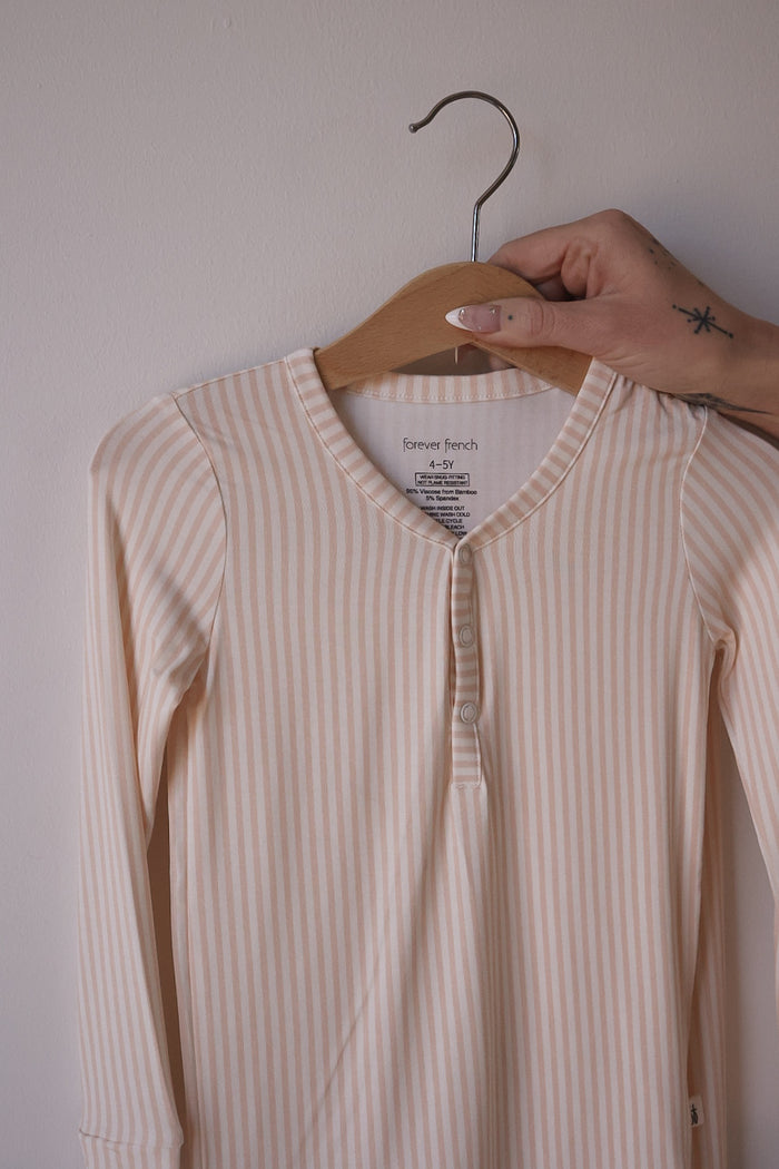 A hand holds a hanger with Bamboo Two Piece Pajamas in Sadie Stripe by forever french baby, featuring long sleeves, cream and light peach stripes, buttons at the collar, and a branded tag, against a plain light wall.