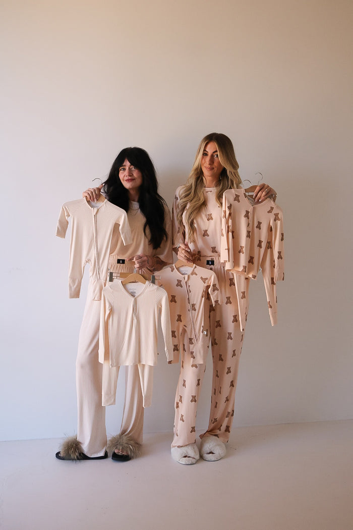 Two women in pajamas and slippers stand against a plain wall, each holding up matching forever french baby Bamboo Two Piece Pajamas | Sadie Stripe—light, neutral-toned bamboo sleepwear that’s gentle on sensitive skin and perfect for cozy nights.