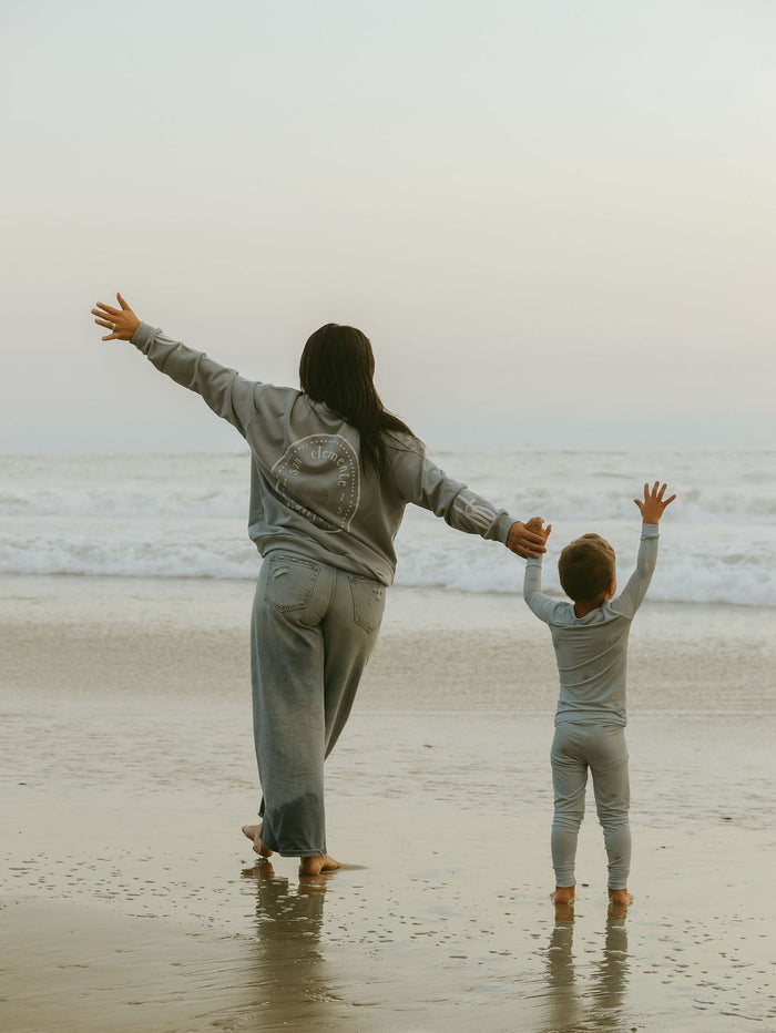 An adult and child in forever french baby's Bamboo Two Piece Pajamas | San Clemente stand barefoot on a beach with arms joyfully raised, overcast sky above and gentle waves rolling in behind them.