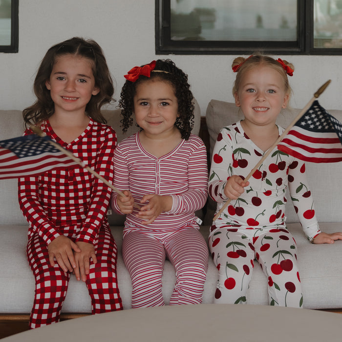 Three young girls in hypo-allergenic forever french baby Bamboo Two Piece Pajamas | Heritage Stripe sit on a couch, smiling. Two hold small American flags, and their festive sleepwear creates a playful, patriotic vibe.