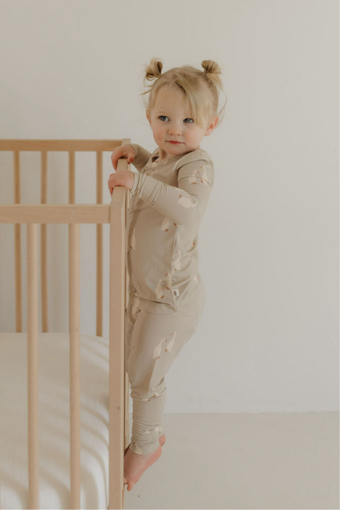 A young child with light hair in two buns stands barefoot by a light wooden crib, wearing forever french baby’s Bamboo Two Piece Pajamas | Silly Goose, featuring a beige bear print, in a minimal white room.