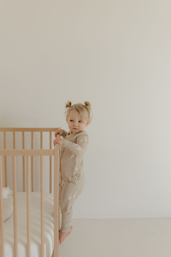 A toddler with two pigtails stands inside a wooden crib, dressed in Silly Goose Bamboo Two Piece Pajamas by forever french baby in soft beige. She clings to the side rail and gazes at the camera against a plain, off-white background.
