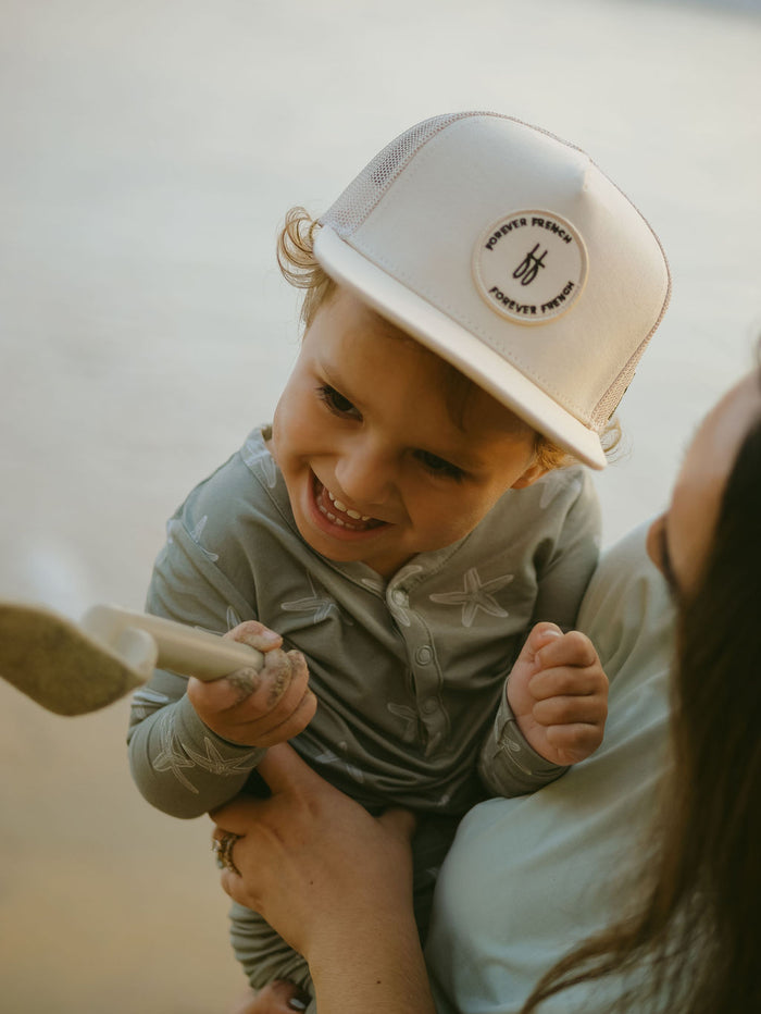 A happy young child wearing the forever french baby Bamboo Two Piece Pajamas | Starfish and a light-colored hat is held by an adult near water and sand, showcasing comfy, hypoallergenic playtime clothing.
