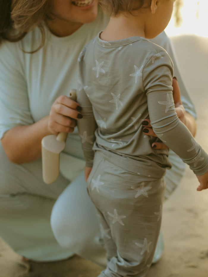 A woman smiles and kneels beside a child in forever french baby Bamboo Two Piece Pajamas | Starfish, holding a small beige toy shovel on sandy ground.