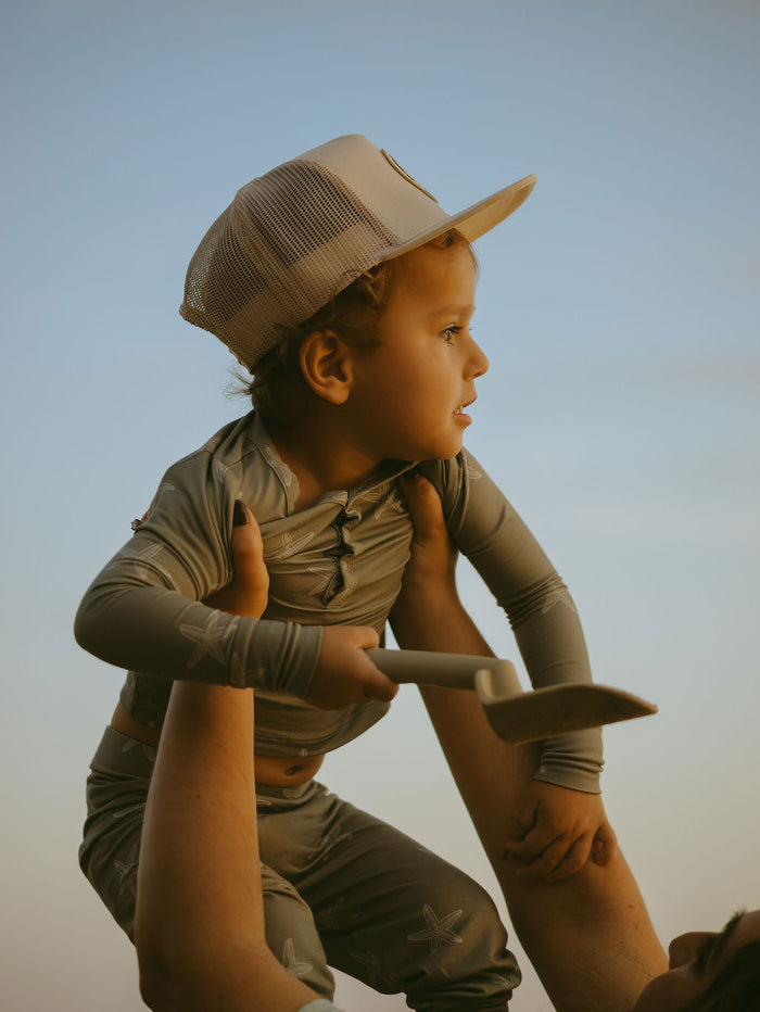 A young child wearing forever french baby's Bamboo Two Piece Pajamas | Starfish and a hat is lifted by an adult, holding a small shovel and gazing sideways against a blue sky.