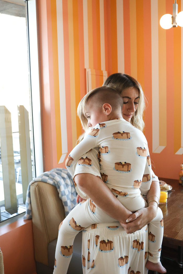 A woman hugs a young child, both wearing matching forever french baby Bamboo Two Piece Pajamas | Sunday Stack with a dog print, in a cozy room with orange and white striped walls and natural light from a window.