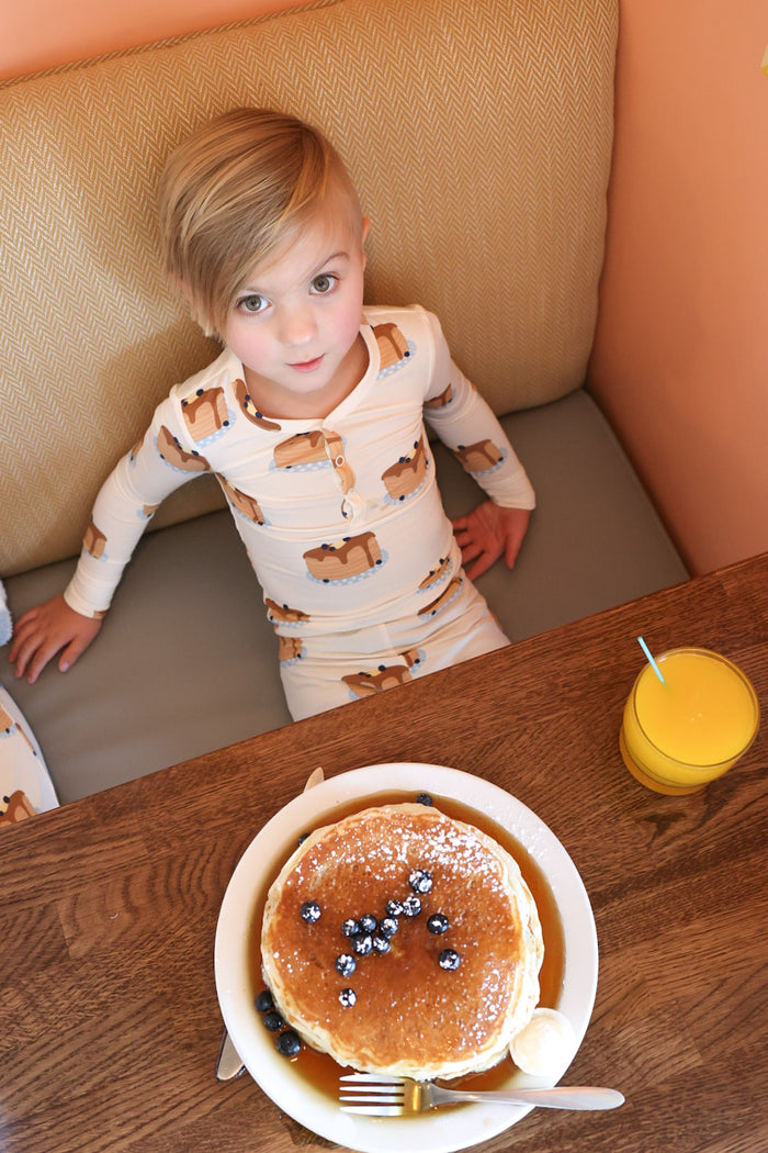 A young child wearing forever french baby’s Bamboo Two Piece Pajamas | Sunday Stack sits at a wooden table with a plate of blueberry pancakes dusted with powdered sugar and a glass of orange juice in front of them.