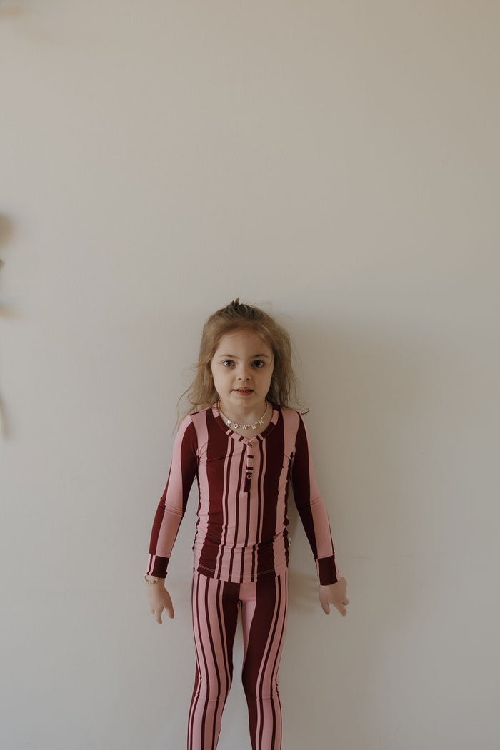 A young child with long brown hair stands against a white wall, wearing forever french baby's Bamboo Two Piece Pajamas | Sweetheart Stripes—cozy maroon and pink striped, hypo-allergenic sleepwear with long sleeves and leggings.