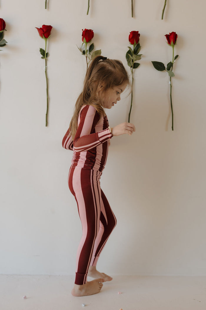 A young child wears forever french baby’s Bamboo Two Piece Pajamas in Sweetheart Stripes, standing sideways in front of a white wall adorned with vertical red roses.