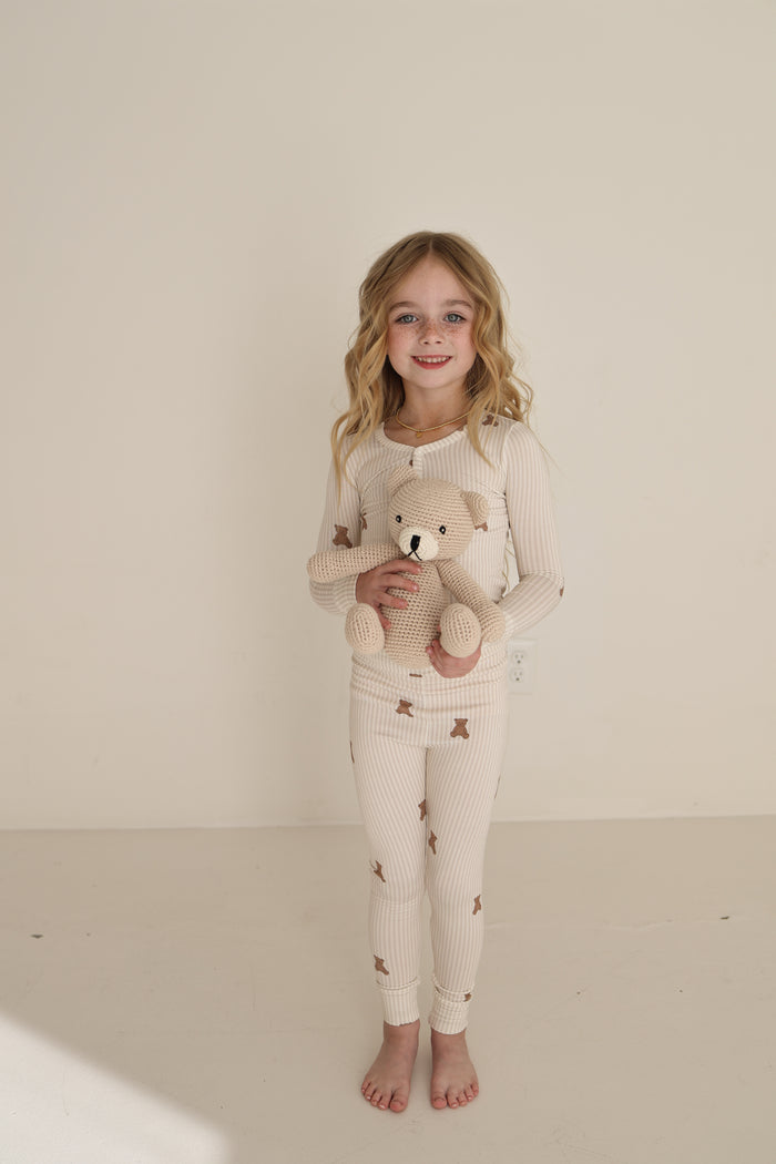 A young girl with long blonde hair smiles barefoot against a light backdrop, holding a knitted teddy, while wearing forever french baby’s Bamboo Two Piece Pajamas | Teddy—cozy, hypo-allergenic sleepwear for kids.