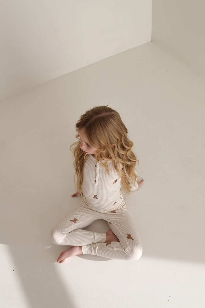 A young child with long, wavy hair sits barefoot on a white floor in forever french baby’s Bamboo Two Piece Pajamas | Teddy, featuring a light-colored star pattern, gazing off to the side as sunlight brightens the nearby floor.