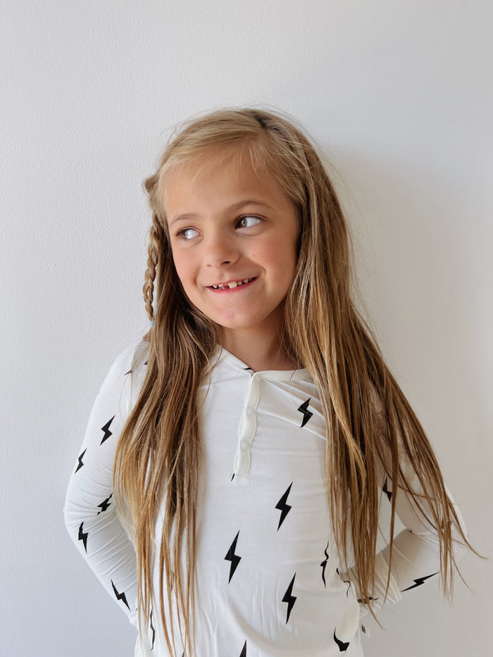 A young girl with long blonde hair smiles while looking to her right. She is wearing hypoallergenic Bamboo Two Piece Pajamas with a Black & White Lightning Bolt design, by forever french baby, and has her hands behind her back. The background is a plain white wall.