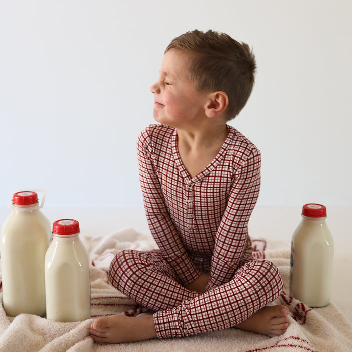 A young boy in forever french baby Bamboo Two Piece Pajamas | Window Pane sits cross-legged on a blanket, smiling with his eyes closed. Three glass bottles of milk with red caps surround him against a plain white background.