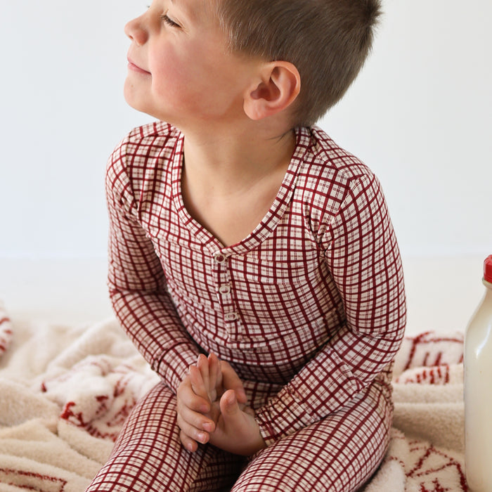 A young child in forever french baby Bamboo Two Piece Pajamas | Window Pane, sits cross-legged on a patterned blanket, smiling off to the side. A bottle is nearby, and the breathable sleepwear adds comfort to the minimal background.