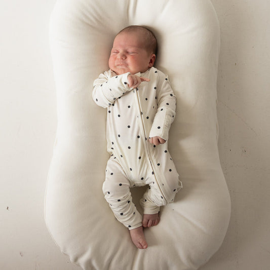 A newborn in forever french baby's Bamboo Zip Pajamas | Annie—white sleepwear with black polka dots—sleeps on a cushioned white lounger against a plain background.