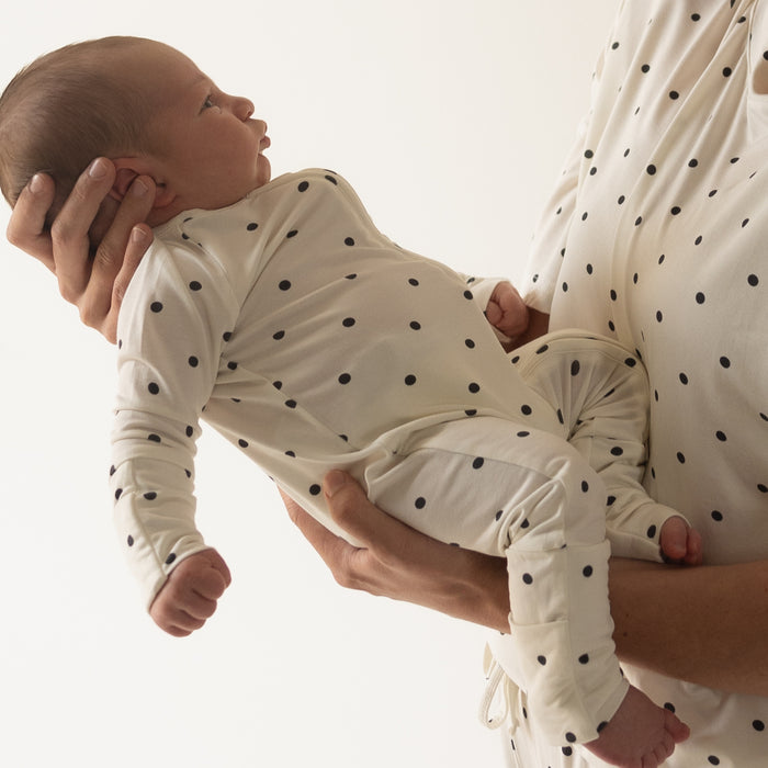 An adult holds a baby dressed in forever french baby's Bamboo Zip Pajamas | Annie. Both wear matching, breathable black polka dot bamboo outfits that stand out against a light background.